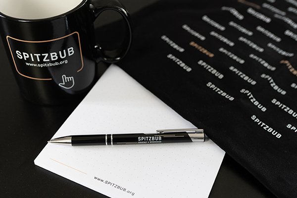 Das versteht man unter Corporate Identity. Dieses Bild zeigt einen Kugelschreiber, eine Tasse, einen Notizblock und eine Tasche von Spitzbub.